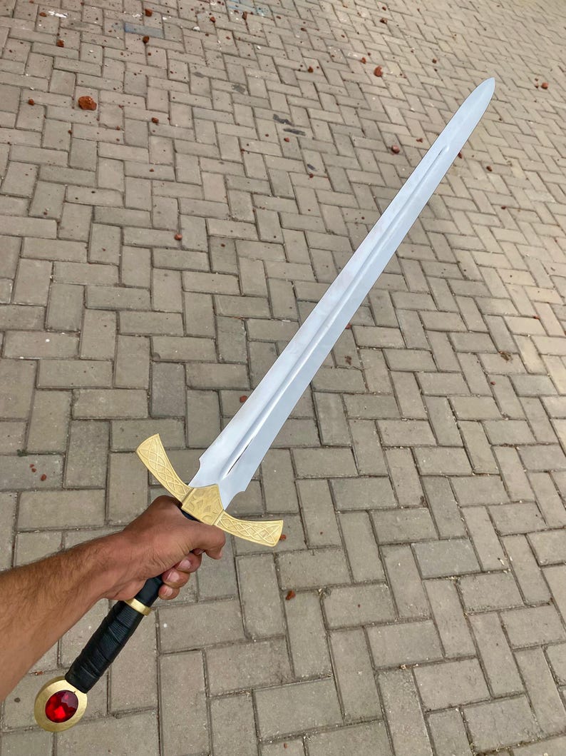 May include: A silver sword with a gold hilt and a red jewel on the pommel. The sword is held by a hand against a background of gray brick.