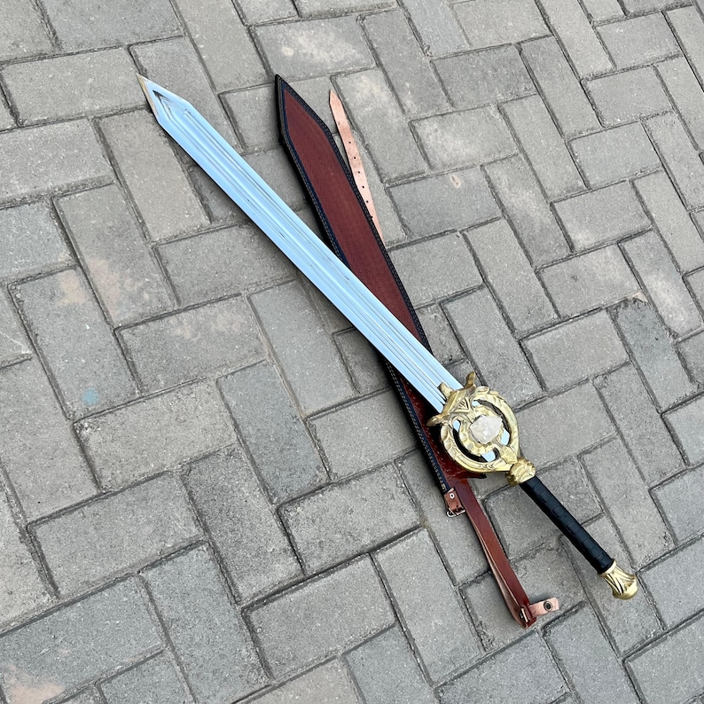 Puede incluir: Una espada decorativa con una hoja azul claro y una empu&ntilde;adura dorada. La espada descansa en una vaina de cuero marr&oacute;n con una correa a juego. La empu&ntilde;adura presenta un dise&ntilde;o circular con una piedra transparente en el centro. El mango es negro.