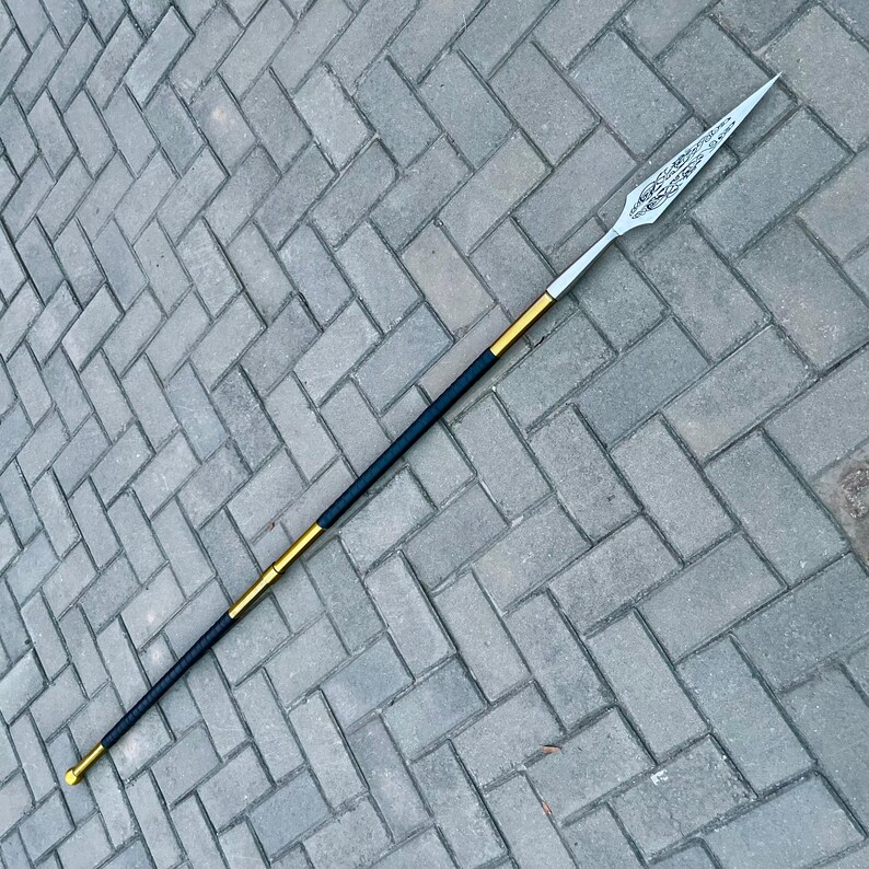 May include: A spear with a silver blade and a black and gold shaft. The blade has an ornate design. The shaft is approximately 6 feet long. The spear is lying on a gray brick surface.