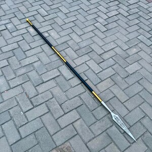May include: A long spear with a silver blade and a black and gold shaft. The spearhead has an ornate design. The shaft is black with gold accents and a gold tip.
