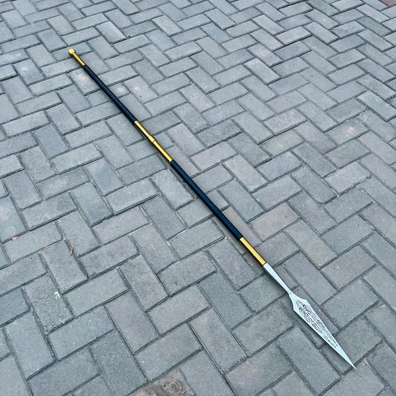 May include: A long spear with a silver blade and a black and gold shaft. The spearhead has an ornate design. The shaft is black with gold accents and a gold tip.