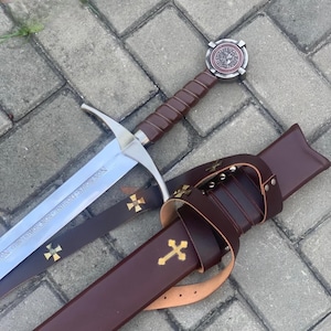 Pode incluir: Uma espada de estilo medieval com uma lâmina prateada e um cabo envolto em couro castanho. A espada apresenta uma cruz decorativa na bainha e no cabo. A bainha é de couro castanho. A lâmina da espada tem a inscrição "NON NOBIS DOMINE NON NOBIS SED NOMINI TUO DA GLORIAM".