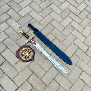May include: A sword, shield, and sheath set. The sword has a silver blade and a brown and gold handle. The shield is burgundy with a gold emblem. The sheath is dark blue. The set is displayed on a brick surface.