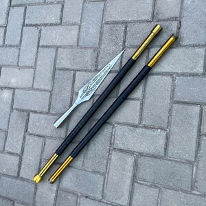 May include: A silver spearhead with an ornate design rests beside two black and gold poles. The poles have a black grip and gold accents on the ends. The items are displayed on a gray brick surface.