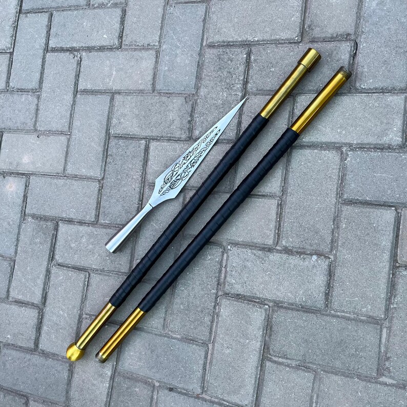 May include: A silver spearhead with an ornate design rests beside two black and gold poles. The poles have a black grip and gold accents on the ends. The items are displayed on a gray brick surface.
