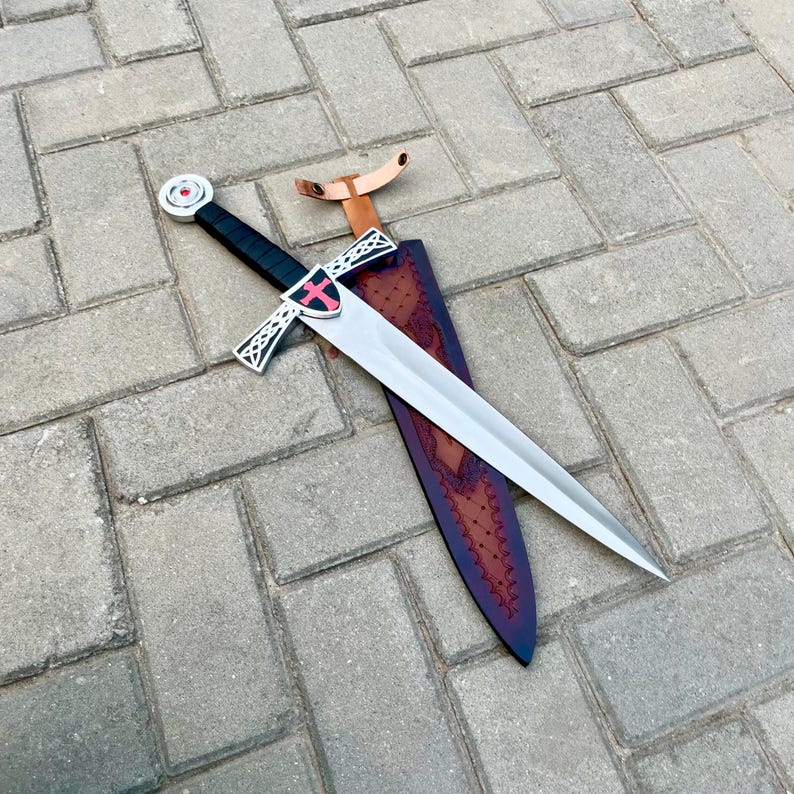 May include: A medieval-style sword with a silver blade and a black handle. The hilt features a red cross on a white shield. The sword rests on a brown leather sheath with decorative stitching. The sword is approximately 30 inches long.