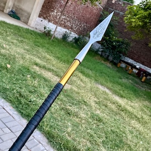 May include: A spear with a silver, engraved blade and a gold and black handle. The spearhead has a decorative design. The handle is wrapped in black material. The spear is set against a background of green grass and a brick wall.