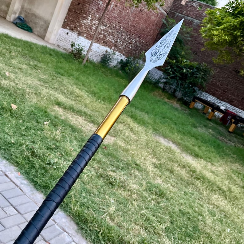 May include: A spear with a silver, engraved blade and a gold and black handle. The spearhead has a decorative design. The handle is wrapped in black material. The spear is set against a background of green grass and a brick wall.