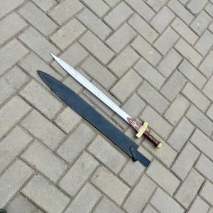 Puede incluir: Una espada plateada con empu&ntilde;adura marr&oacute;n y funda negra. La espada tiene un s&iacute;mbolo de tridente cerca de la empu&ntilde;adura. La funda es de cuero negro. La espada est&aacute; sobre una superficie de ladrillos grises.