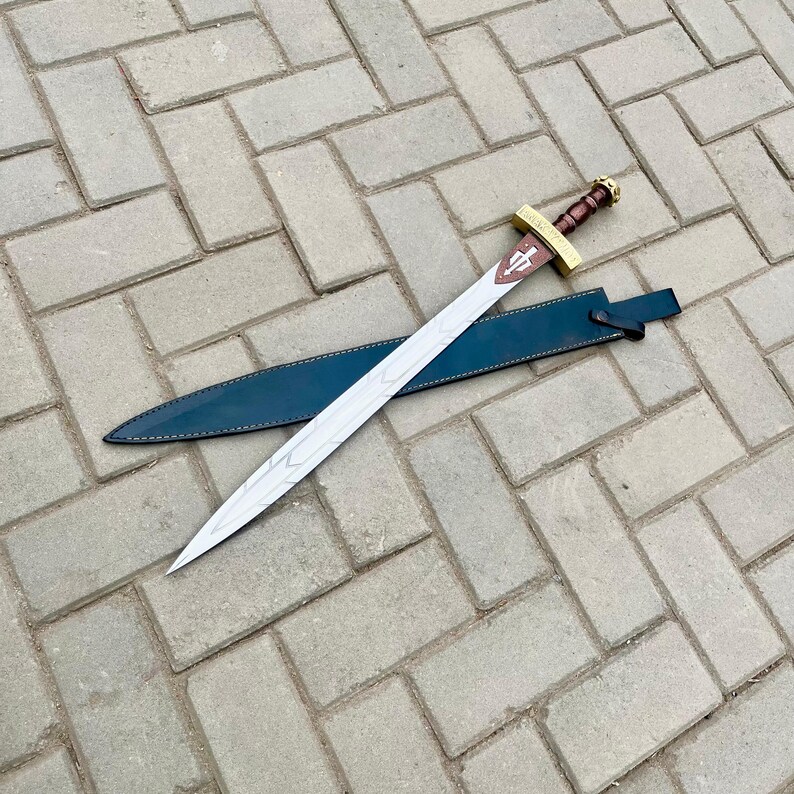 Puede incluir: Una espada de hoja plateada con empu&ntilde;adura marr&oacute;n y detalles dorados, sobre una funda de cuero azul oscuro. La espada presenta un dise&ntilde;o de cruz en la guarda. La hoja tiene un patr&oacute;n grabado decorativo.