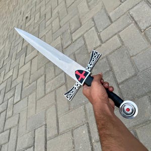 May include: A silver-bladed sword with a black and white crossguard and a red cross emblem. The handle is black with a silver pommel and a red gem. The sword is held against a gray brick background.