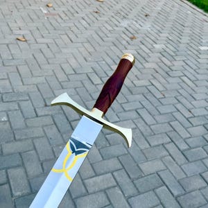 May include: A long sword with a polished silver blade and a gold-colored crossguard. The handle is dark brown with gold accents. The blade features a yellow and black design.