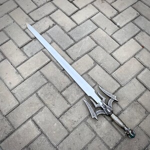 May include: A silver sword with an elaborate handle and a long, straight blade. The hilt has detailed patterns and a decorative pommel. The sword is positioned on a grey brick surface.