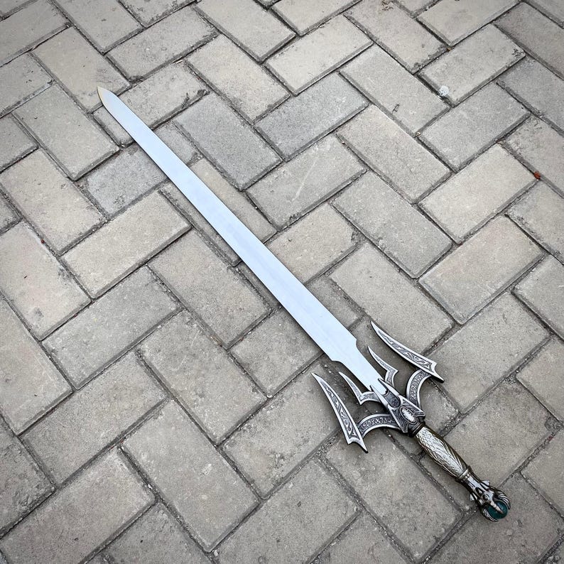 May include: A silver sword with an elaborate handle and a long, straight blade. The hilt has detailed patterns and a decorative pommel. The sword is positioned on a grey brick surface.