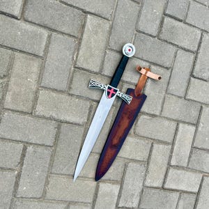 May include: A medieval-style sword with a silver blade, black handle, and a red cross emblem on the hilt. The sword rests on a brown leather sheath with a light brown leather strap. The sword is approximately 30 inches long.