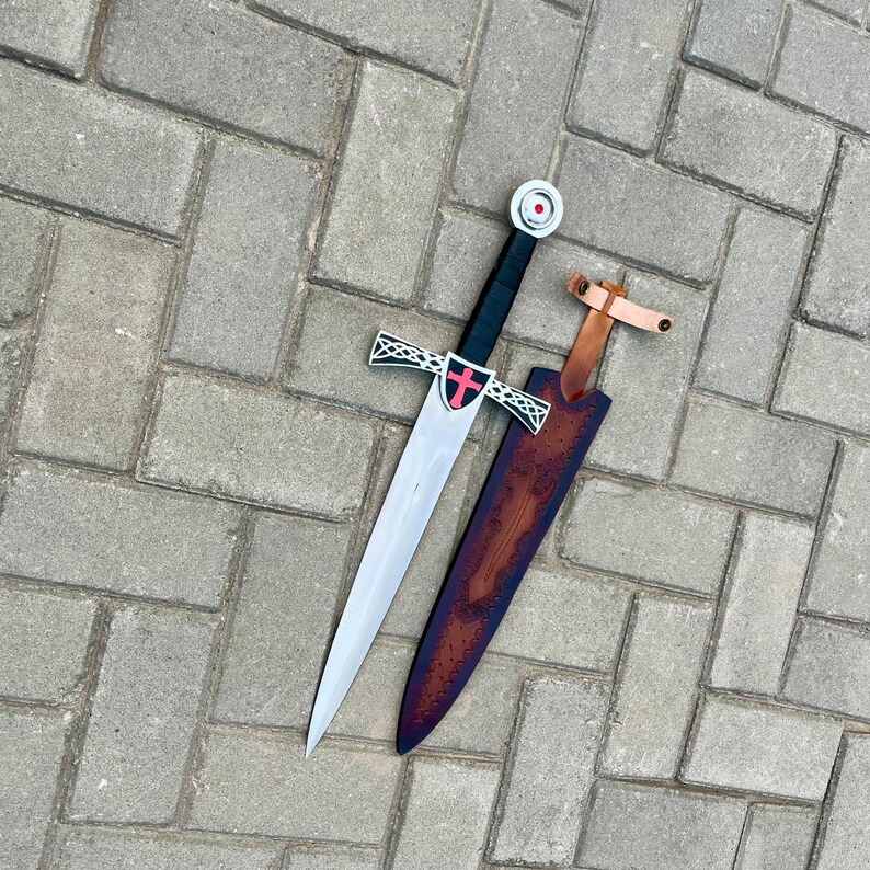 May include: A medieval-style sword with a silver blade, black handle, and a red cross emblem on the hilt. The sword rests on a brown leather sheath with a light brown leather strap. The sword is approximately 30 inches long.