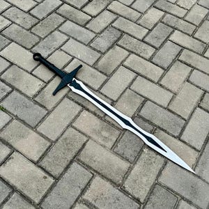以下が含まれることがあります： 黒と銀の剣で、ガードと尖った刃が付いています。 剣の柄は黒で、刃は白い中心に黒の装飾要素があります。 剣は灰色のレンガの表面に置かれています。