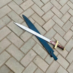 Puede incluir: Una espada plateada y marr&oacute;n con una funda de cuero. La espada tiene una empu&ntilde;adura decorativa y una hoja con un dise&ntilde;o detallado. La funda es azul oscuro y la espada est&aacute; sobre una superficie de ladrillo.