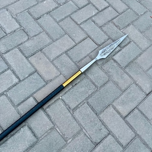 May include: A spear with a silver blade featuring an etched design. The shaft is black with a gold band near the blade. The spear is lying on a gray brick surface.