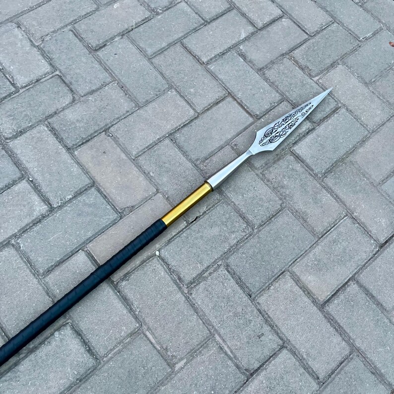 May include: A spear with a silver blade featuring an etched design. The shaft is black with a gold band near the blade. The spear is lying on a gray brick surface.