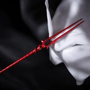 May include: A red spear with a spiraled shaft and a forked tip. The spear is made of metal and has a glossy finish. The background is a soft white fabric and a dark, blurred area.