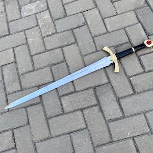 May include: A silver sword with a black leather grip and a gold pommel with a red jewel. The sword is lying on a gray brick surface.