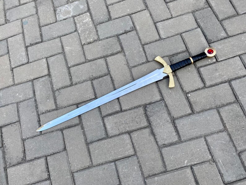 May include: A silver sword with a black leather grip and a gold pommel with a red jewel. The sword is lying on a gray brick surface.