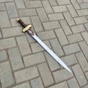 Puede incluir: Una espada decorativa con una hoja plateada y un mango marr&oacute;n. La guarda es dorada con un agarre marr&oacute;n. La hoja tiene un patr&oacute;n decorativo y un emblema granate. La espada est&aacute; sobre una superficie de ladrillos grises.