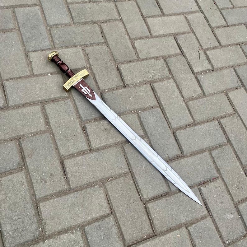 Puede incluir: Una espada decorativa con una hoja plateada y un mango marr&oacute;n. La guarda es dorada con un agarre marr&oacute;n. La hoja tiene un patr&oacute;n decorativo y un emblema granate. La espada est&aacute; sobre una superficie de ladrillos grises.