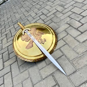 Könnte beinhalten: Ein goldener Schild mit einem Bronze-Adler-Emblem und ein langes, silbernes Schwert mit einem dekorativen Knauf. Schild und Schwert liegen auf einer grauen Ziegeloberfläche. Das Schwert liegt diagonal über dem Schild.