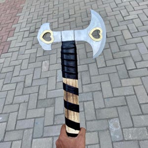 May include: A silver double-headed axe with a black and brown wooden handle. The axe has two heart-shaped cutouts on each side of the blade.