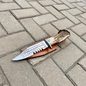 May include: A handcrafted hunting knife with a polished steel blade and a serrated edge. The handle is made of antler, and the knife rests in a brown leather sheath. The blade features etched text.