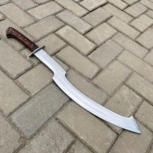 May include: A curved, silver-colored sword with a brown wooden handle. The sword is lying on a gray brick surface.