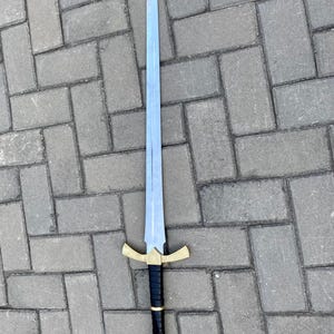 May include: A silver sword with a black leather grip and a gold pommel with a red jewel. The sword has a cross guard with a gold finish.