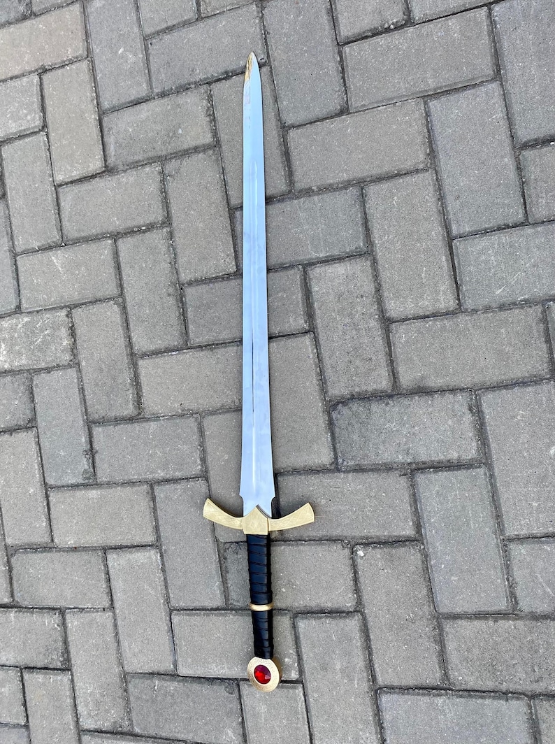 May include: A silver sword with a black leather grip and a gold pommel with a red jewel. The sword has a cross guard with a gold finish.