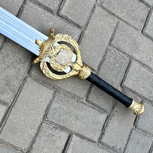 Puede incluir: Una espada de fantasía con una hoja plateada y una empuñadura dorada ornamentada. La empuñadura presenta una piedra central y un agarre negro texturizado. El diseño de la espada es detallado y parece inspirado en temas medievales o fantásticos.