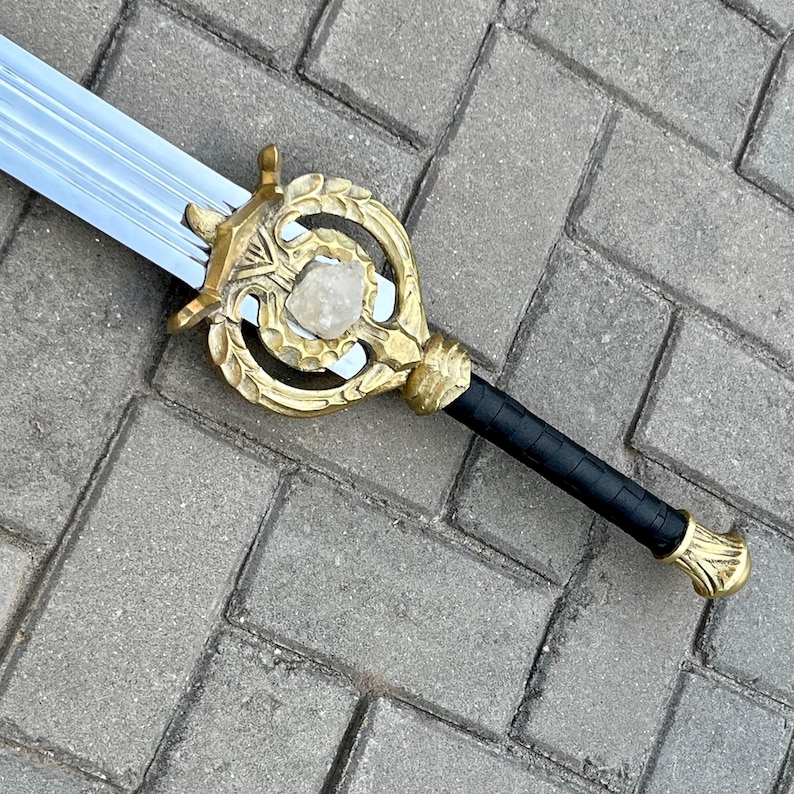 Puede incluir: Una espada de fantas&iacute;a con una hoja plateada y una empu&ntilde;adura dorada ornamentada. La empu&ntilde;adura presenta una piedra central y un agarre negro texturizado. El dise&ntilde;o de la espada es detallado y parece inspirado en temas medievales o fant&aacute;sticos.
