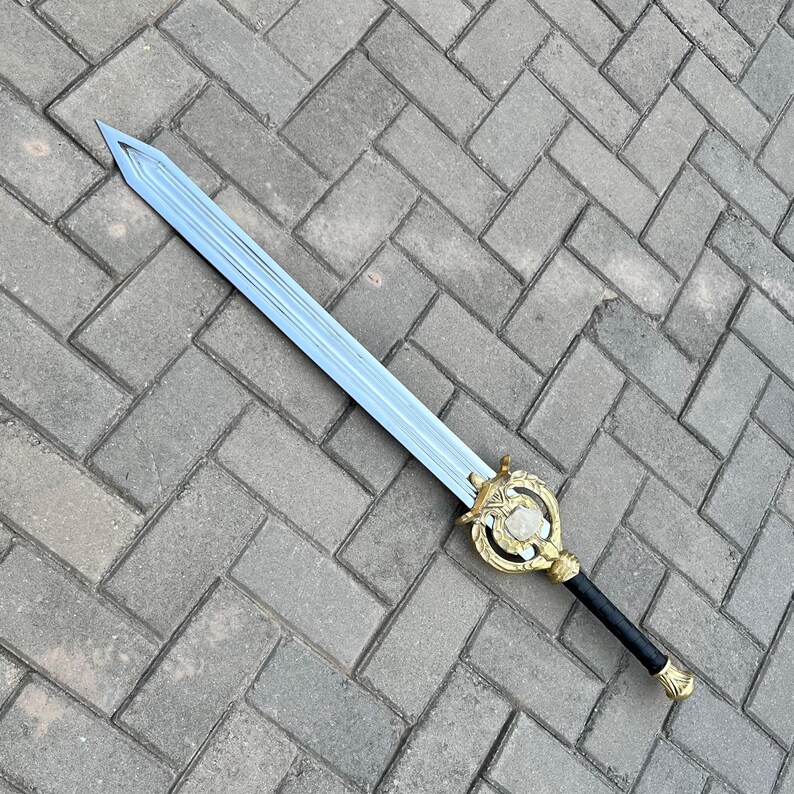 Puede incluir: Una espada de hoja plateada con una empu&ntilde;adura dorada decorativa y un mango negro. La empu&ntilde;adura presenta un dise&ntilde;o ornamentado con una piedra blanca central. La espada est&aacute; sobre una superficie de ladrillos grises. Un art&iacute;culo de fantas&iacute;a.