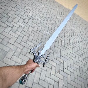 May include: A silver fantasy sword with an ornate hilt and a long, straight blade. The hilt features intricate designs and a green gem at the base. The sword is held against a grey brick background.