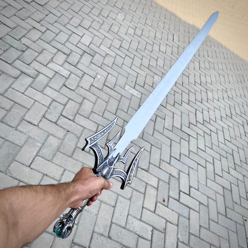 May include: A silver fantasy sword with an ornate hilt and a long, straight blade. The hilt features intricate designs and a green gem at the base. The sword is held against a grey brick background.