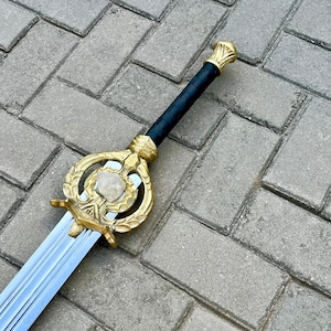 Puede incluir: Una espada decorativa con empu&ntilde;adura y guarda doradas, con una piedra central. El mango est&aacute; envuelto en material negro. La hoja es plateada y tiene un dise&ntilde;o estriado. La espada se exhibe sobre una superficie de ladrillo gris.