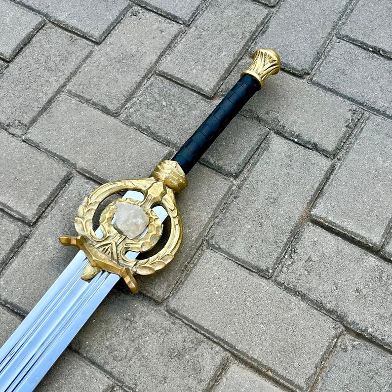 Puede incluir: Una espada decorativa con empu&ntilde;adura y guarda doradas, con una piedra central. El mango est&aacute; envuelto en material negro. La hoja es plateada y tiene un dise&ntilde;o estriado. La espada se exhibe sobre una superficie de ladrillo gris.