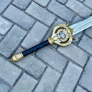 Puede incluir: Una espada decorativa con una empuñadura dorada y un mango negro. La empuñadura presenta un diseño ornamentado con una piedra central. La hoja es plateada y tiene un diseño estriado. La espada está sobre una superficie de ladrillos grises.