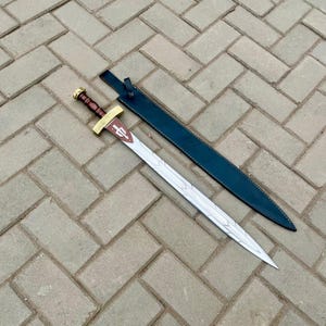 Puede incluir: Una espada decorativa con una hoja plateada y un mango marr&oacute;n. La empu&ntilde;adura es dorada con un dise&ntilde;o decorativo. La espada va acompa&ntilde;ada de una funda azul oscuro. La espada est&aacute; sobre una superficie de ladrillo.