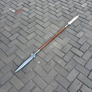 Może przedstawiać: Dwustronna włócznia z dwoma ostrymi, metalowymi ostrzami na każdym końcu. Środkowy trzon owinięty jest teksturowanym materiałem w jasnobrązowym kolorze, zapewniającym uchwyt pomiędzy dwoma metalowymi grotami wystającymi ze środka.