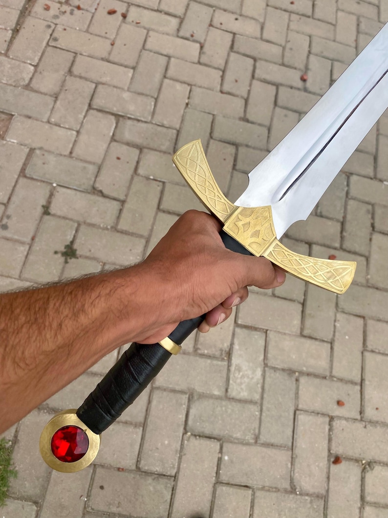 May include: A sword with a gold hilt and a red jewel at the pommel. The blade is silver and the hilt has intricate designs.