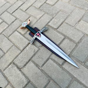 May include: A silver sword with a black handle and a red cross emblem on the guard. The sword is displayed on a wooden sheath with a leather strap. The blade is sharp and pointed.