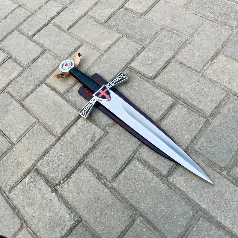 May include: A silver sword with a black handle and a red cross emblem on the guard. The sword is displayed on a wooden sheath with a leather strap. The blade is sharp and pointed.