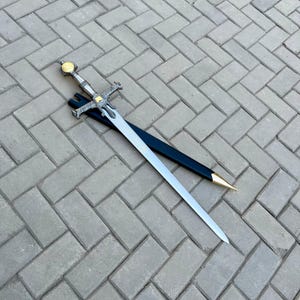 May include: A decorative sword with a silver blade and ornate gold and silver hilt, resting on a gray brick surface. The sword is accompanied by a black sheath with a gold-colored tip. The blade is long and sharp.