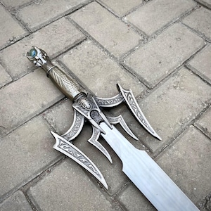 May include: An intricately designed silver fantasy sword. The handle has a dragon-like motif with a blue gem. The sword features decorative, curved crossguards and a wide, flat blade. The sword is lying on a grey brick surface.
